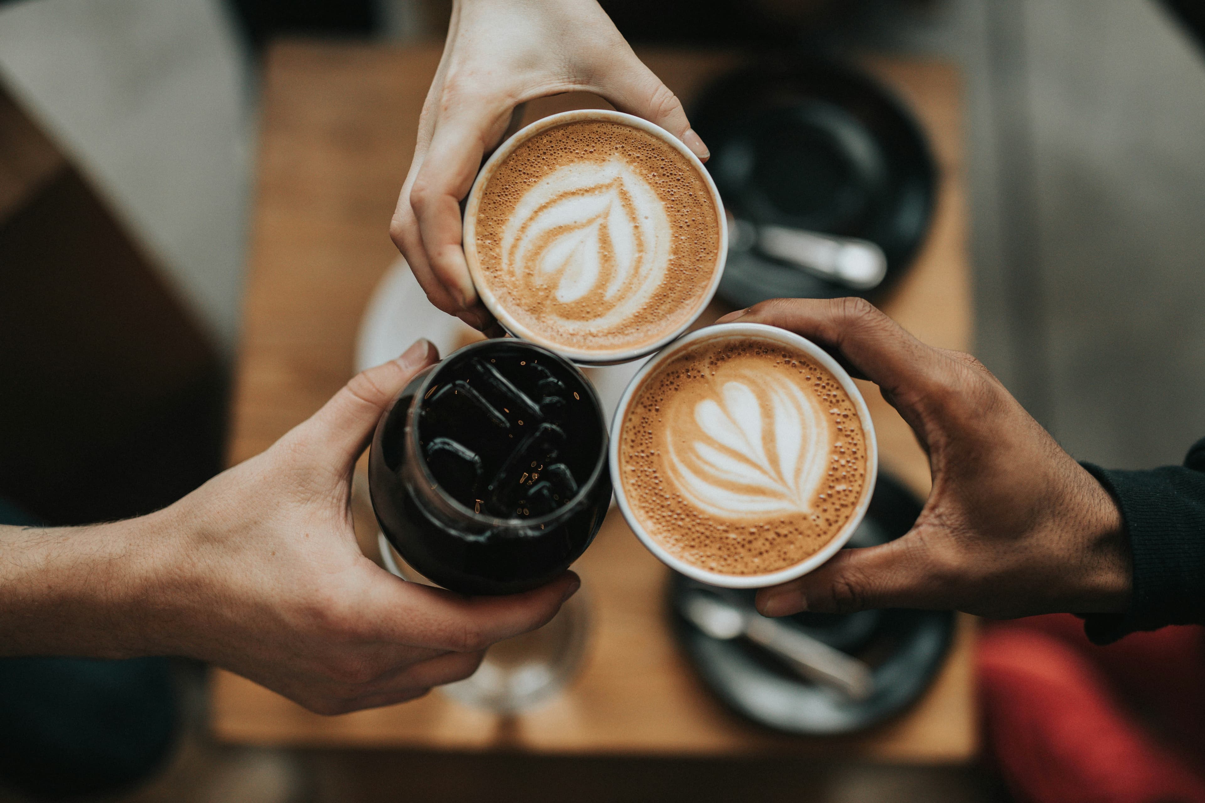 Friends raising two lattes and an iced coffee over a café table